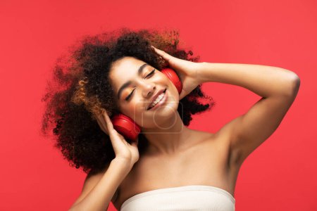Foto de People, music, emotions concept. Young afro american female with dances in rhythm of melody, closes eyes listens loud song in headphones over red background. - Imagen libre de derechos