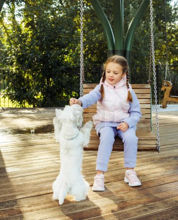 Foto de A seven-year-old girl swings on a swing and plays with a dog in a summer park.Animal friendship and happy childhood concept. - Imagen libre de derechos