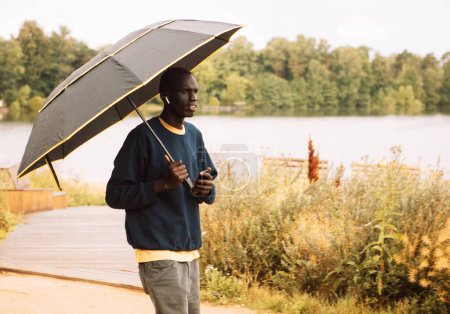 Téléchargez les photos : Un jeune Afro-Américain vêtu de vêtements décontractés se promène par une matinée nuageuse sous la pluie près d'un lac. Il tient un parapluie. Concept de style de vie. - en image libre de droit