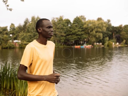 Photo for Cheerful black man wearing yellow t-shirt jogging outdoor along river bank early in morning. Sport and lifestyle concept. - Royalty Free Image
