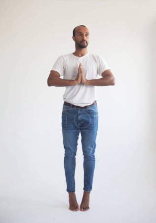 Foto de Joven afro hombre vistiendo camiseta blanca, concentrando su mente, manteniendo las manos namaste gesto, meditando, yoga ejercicio respiración técnica reducir el estrés. - Imagen libre de derechos