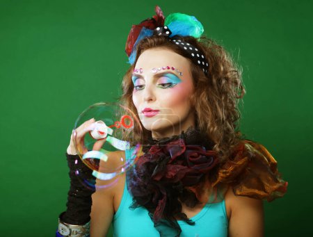Téléchargez les photos : Young curly woman with creative make-up blowing soap bubbles over green background. Doll style. - en image libre de droit