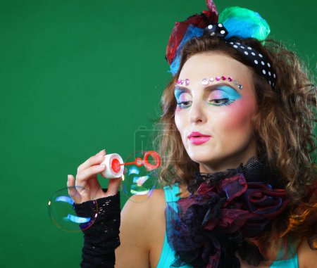 Photo for Young curly woman with creative make-up blowing soap bubbles over green background. Doll style. - Royalty Free Image