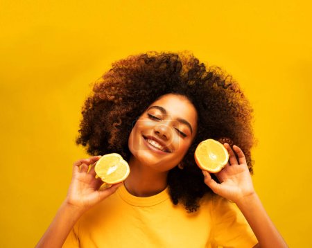Foto de Lifestyle, food, diet and people concept: Charming dark-skinned young woman in a yellow t-shirt holds orange slices. Beautiful model posing with her eyes closed, on a yellow studio background. - Imagen libre de derechos