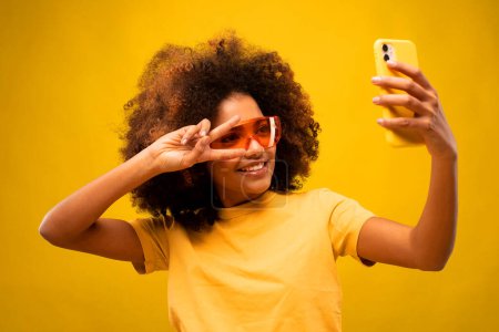 Téléchargez les photos : Style de vie, tehnology et concept de personnes : Photo de jeune femme africaine heureux sourire positif montrer la paix v-signe faire selfie smartphone isolé sur fond de couleur jaune - en image libre de droit
