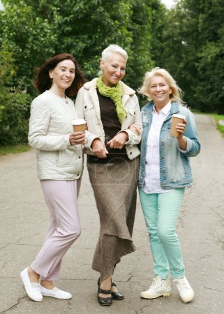 Téléchargez les photos : Lifestyle, emotion, old people and gold age concept. Three elderly ladies in casual clothes are walking in the park. Coffee and socializing, friendship in retirement. - en image libre de droit