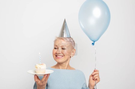 Téléchargez les photos : People, party, aging and maturity concept. Beauitful middle aged female wearing conical hat celebrating birthday, posing isolated with cupcake with candle and blue balloon in her hands. Happy time. - en image libre de droit