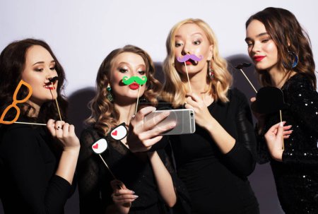 Photo for Celebration, party and emotions concept: four charming ladies dressed in black cocktail dresses with party falce mustache take selfie. - Royalty Free Image