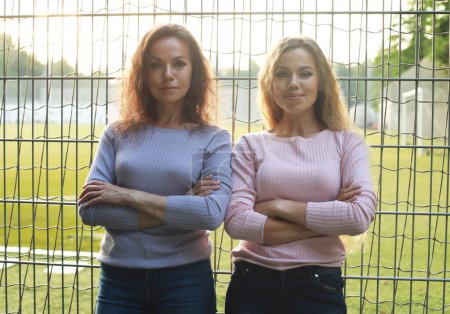 Téléchargez les photos : Two women with long curly hair stand with their arms crossed next to a football field. Adult mother and her beautiful young daughter. Two generations. - en image libre de droit