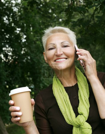 Téléchargez les photos : Beautiful elderly woman with short blond hair drinks coffee and chats on the phone in a summer park. Lifestyle and old people concept. - en image libre de droit