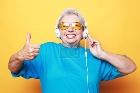Téléchargez les photos : Elderly woman listening to music and show thumbs up over yellow background. Lifestyle and old people concept. - en image libre de droit
