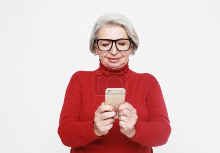 Téléchargez les photos : Tehnology and old people concept: Pretty senior female in casual outfit using modern smartphone and laughing out loud while standing on white background. Old woman watching funny videos on internet. - en image libre de droit