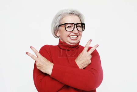 Photo for Portrait of funny senior gray-haired woman wearing glasses and red sweater doing a rock and roll symbol over white background. Lifestyle concept. - Royalty Free Image