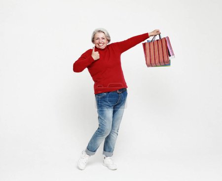 Foto de Foto de longitud completa de positivo alegre gris pelo blanco anciano mujer muestran signo ok disfrutar del tiempo libre compras mantenga bolsas usan pantalones vaqueros y suéter rojo aislado sobre fondo blanco - Imagen libre de derechos