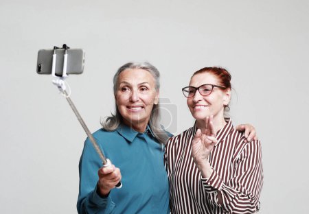 Photo for Two happy elderly women frineds taking a selfie with a stick isolated on grey background - Royalty Free Image