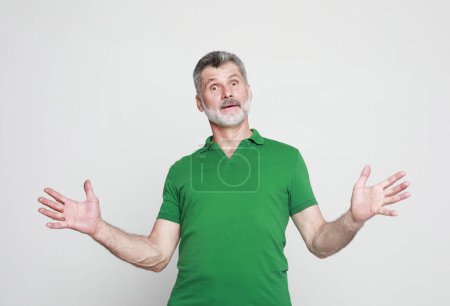 Foto de Surprised joyful elderly man wearing green t-shirt with a gray beard raises his hands in amazement. - Imagen libre de derechos