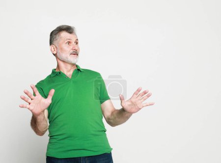 Photo for Surprised joyful elderly man wearing green t-shirt with a gray beard raises his hands in amazement. - Royalty Free Image
