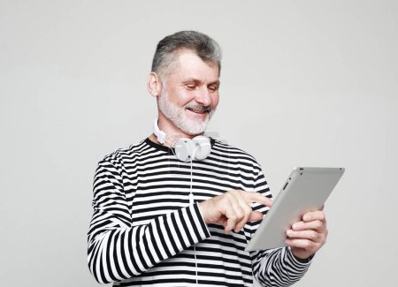 Foto de Charming elderly man communicates with friends using a digital tablet and headphones. Portrait over grey background. - Imagen libre de derechos