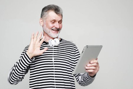 Photo for Charming elderly man communicates with friends using a digital tablet and headphones. Portrait over grey background. - Royalty Free Image