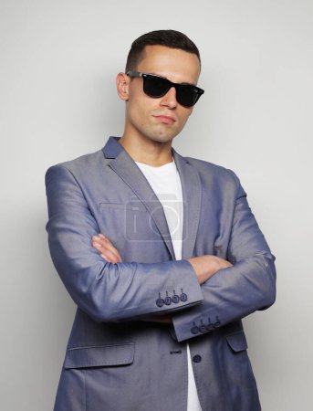 Foto de Waist-up shot of young man wearing sunglasses looking at camera with crossed arms over grey background - Imagen libre de derechos