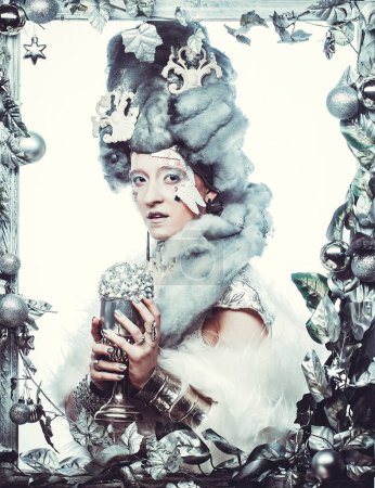 Foto de Concepto de fiesta, Navidad y gente: Mujer joven en imagen creativa con maquillaje artístico plateado sosteniendo la bola de Navidad, posando en un marco plateado decorado con decoraciones navideñas. Retrato de invierno. - Imagen libre de derechos
