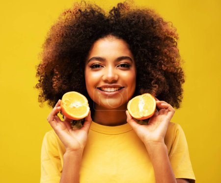 Foto de Estilo de vida, comida, dieta y concepto de personas: Foto de una joven afroamericana que sostiene rebanadas de naranja se ven sorprendidas aisladas sobre un fondo de color amarillo. - Imagen libre de derechos