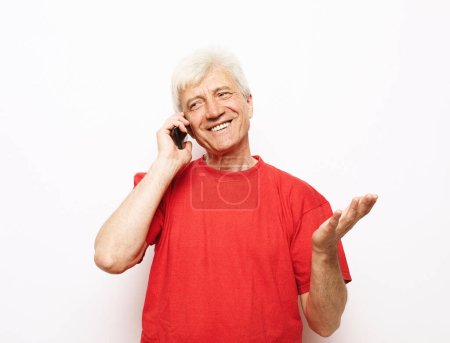 Téléchargez les photos : Modern lifestyle, tehnology and old people concept:old happy man dressed in a red t-shirt holds a smartphone and hears good news. Pensioner smiles and rejoices while standing on a white background.. - en image libre de droit
