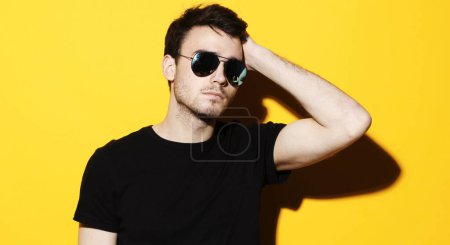 Photo for Lifestyle, fashion and people concept: Portrait of a young handsome man dressed casual and wearing sunglasses on yellow background - Royalty Free Image