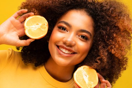 Téléchargez les photos : Healthy summer, diet and people concept: Cheerful afro woman holding oranges near ears, yellow background - en image libre de droit