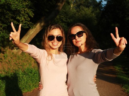 Téléchargez les photos : Beautiful mother and adult daughter, two generations. Summer day in the park. They pose and show the sign of victory. Sunset time, golden hour. - en image libre de droit