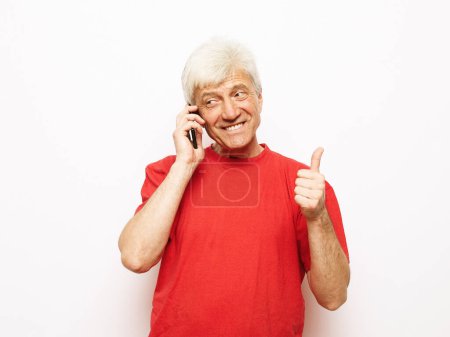 Téléchargez les photos : Style de vie moderne, tehnologie et concept de personnes âgées : Un homme âgé vêtu d'un t-shirt rouge tient un smartphone et entend de bonnes nouvelles. Pensionné sourit et se réjouit en se tenant debout sur un fond blanc.. - en image libre de droit
