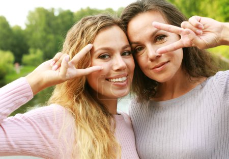 Photo for Lifestyle, family and people concept: mature mother and adult daughter are doing selfie by mobile phone in summer park. Women show v-sign and smiling, - Royalty Free Image