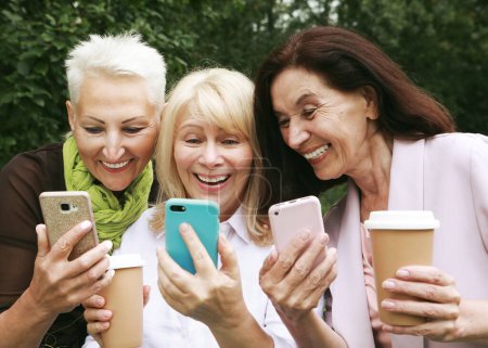 Foto de Three elderly women are smiling and looking photos at the at screen of the phones in the park on a summer day. Golden age. - Imagen libre de derechos