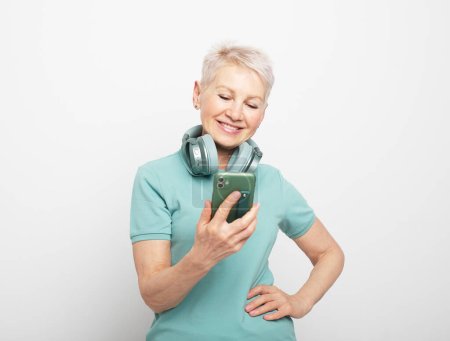 Photo for Elderly woman in headphones reads sms on smartphone over grey background - Royalty Free Image