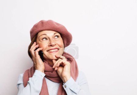 Photo for Modern lifestyle, old people, communication and electronic gadgets concept. Elderly woman wearing pink scarf and hat with smartphone over white grey background. - Royalty Free Image