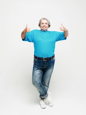 Photo for Lifestyle, emotion and people concept: Elderly happy female wearing jeans and blue shirt giving a thumb up and looking at the camera. Portrait full length. - Royalty Free Image