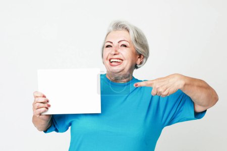 Foto de Viejos y concepto de estilo de vida: Una anciana feliz y encantadora sostiene una hoja de papel en blanco en sus manos y la señala con su dedo. - Imagen libre de derechos