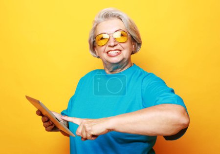 Foto de Education, old people and tehnology concept: Grey haired old smiling woman wearing glasses, reading browsing using holding tablet. Isolated over yellow background. - Imagen libre de derechos