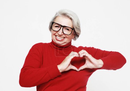 Foto de Perfect, nice, aged, old woman showing heart figure with fingers, looking at camera, celebrating woman's day, standing over white background - Imagen libre de derechos