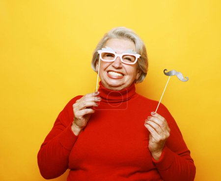 Téléchargez les photos : Lifestyle and old people concept: funny grandmother in red sweater with fake mustache and glasses, laughs and prepares for party over yellow background - en image libre de droit