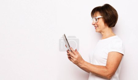 Photo for Old smiling business woman wearing glasses, reading browsing using holding tablet. Isolated over white background - Royalty Free Image