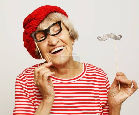 Téléchargez les photos : Style de vie et concept de personnes : grand-mère drôle avec fausse moustache et lunettes, rit et se prépare pour la fête sur fond gris - en image libre de droit