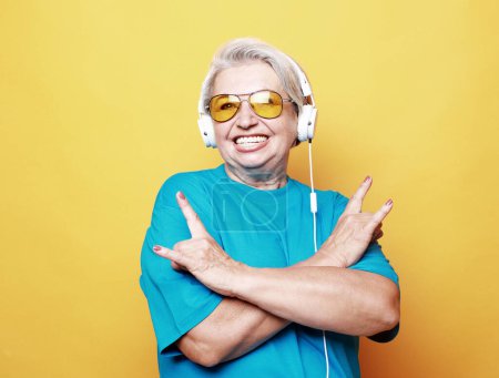 Téléchargez les photos : Amoureux de musique. Portrait d'une femme mûre souriante dans un casque écoutant ses chansons préférées et montrant un signe rock, fond jaune, - en image libre de droit