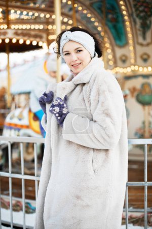Photo for A beautiful young brunette woman in a faux fur coat and snake clothes poses next to a carousel at a Christmas market. - Royalty Free Image