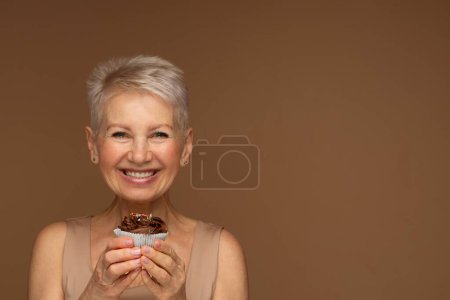 Foto de Una anciana con el pelo corto tiene una magdalena de chocolate en sus manos. Retrato sobre fondo marrón. - Imagen libre de derechos