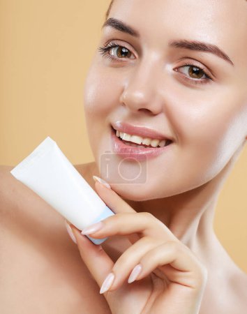 Téléchargez les photos : Beauty Youth Skin Care Concept - Beautiful Caucasian Woman Face Portrait holding and presenting cream tube product. Beautiful Spa model female with Perfect Fresh Clean Skin over beige background. - en image libre de droit