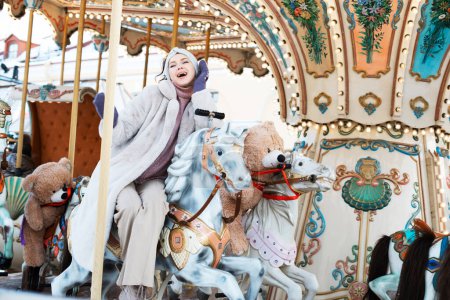 Téléchargez les photos : Cheerful young woman in a faux fur coat and mittens rides a carousel and has fun. Winter happy. - en image libre de droit