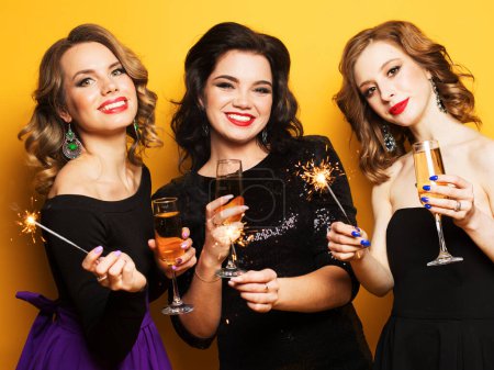 Photo for Portrait of beautiful three young women with a champagne glasses and sparklers over yellow background - Royalty Free Image