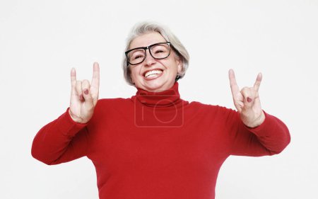 Foto de Retrato de una divertida mujer de pelo gris con gafas y suéter rojo haciendo un símbolo de rock and roll sobre fondo blanco. Concepto de estilo de vida. - Imagen libre de derechos