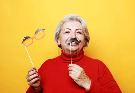 Téléchargez les photos : Lifestyle and old people concept: funny grandmother in red sweater with fake mustache and glasses, laughs and prepares for party over yellow background - en image libre de droit